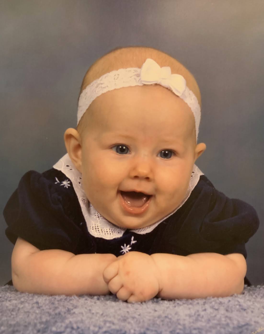 Abby as a baby, smiling in a navy velvet dress with a white bow headband